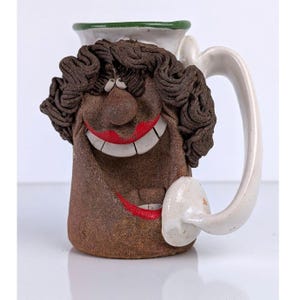 May include: A whimsical ceramic mug with a smiling face design. The mug features a brown face with dark brown curly hair, red lips, and a white handle. The rim of the mug is green.