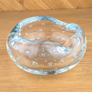 May include: A clear, round ashtray with a light blue rim and numerous air bubbles embedded in the glass. The ashtray sits on a wooden surface, showcasing its unique, handcrafted design. This decorative piece adds a touch of elegance.