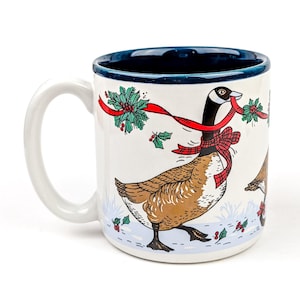 May include: White ceramic mug with a blue rim and handle. The mug features a festive design of a goose wearing a red scarf and carrying holly. The design is set against a white background with a blue base, creating a holiday theme.