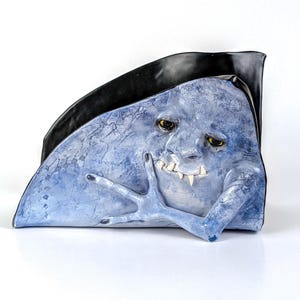 May include: A blue, textured sculpture of a creature with a face, fangs, and a hand touching its face. The sculpture has yellow eyes and black fingernails. A black, curved shape is behind the creature.