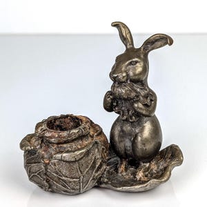 May include: A silver-toned metal figurine of a rabbit holding its paws near its face, next to a small, textured container. The rabbit sits on a leaf-shaped base, and the container has a rough, organic appearance.