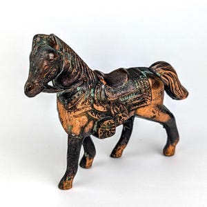 May include: A bronze-colored horse figurine with a saddle. The horse has a detailed mane and tail, with a weathered, aged appearance. The saddle is intricately designed with floral patterns and decorative elements. The figurine is approximately 6 inches long.
