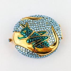 May include: An oval-shaped, gold-toned compact with a turquoise and emerald green dragon design. The compact is encrusted with light blue rhinestones. The dragon is emerald green with turquoise accents.
