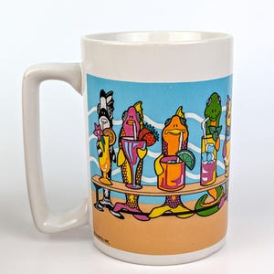 May include: White ceramic mug featuring a colorful illustration of anthropomorphic fish enjoying cocktails at a beach bar. The design includes a blue sky, sandy beach, and various tropical drinks. The mug has a large handle.