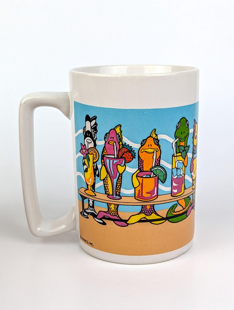 May include: White ceramic mug featuring a colorful illustration of anthropomorphic fish enjoying cocktails at a beach bar. The design includes a blue sky, sandy beach, and various tropical drinks. The mug has a large handle.