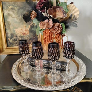 May include: Four vintage wine glasses with dark red bowls and clear stems sit on a silver tray. The glasses have a cut pattern design. A framed painting and a floral arrangement with dried flowers are in the background.