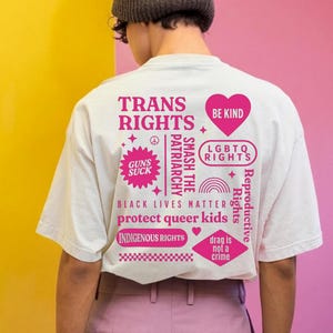 May include: A white t-shirt with pink text and graphics, including "TRANS RIGHTS," "BE KIND," and "BLACK LIVES MATTER." The shirt features various slogans and symbols related to social justice and LGBTQ+ rights. The person is wearing a grey beanie and pink trousers.