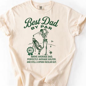 May include: A cream-colored short-sleeve t-shirt featuring a dark green graphic of a skeleton playing golf. The text reads: Best Dad By Par, Ranked No. 1 By Family, Above Average Dad. Perfectly Average Golfer. And Still A Spook-Tacular Guy.