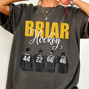 May include: Dark gray t-shirt with the words "BRIAR Hockey" in yellow and white. Below the text are four figures wearing black jerseys with the names and numbers: Graham 44, Logan 22, Di Laurentis 66, and Tucker 46.