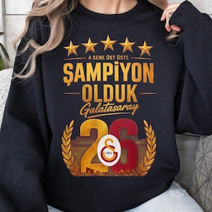 Puede incluir: Sudadera negra con texto dorado y detalles de estrellas. El texto dice "4 SENE &Uuml;ST &Uuml;STE ŞAMPİYON OLDUK Galatasaray 26". El n&uacute;mero 26 es dorado y rojo con un dise&ntilde;o de corona de laurel. La sudadera es de estilo casual y c&oacute;modo.