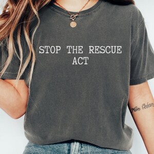 Peut inclure: T-shirt gris foncé avec le texte blanc « STOP THE RESCUE ACT ». Le t-shirt est de style col rond et semble être fait d'une matière douce et confortable. Le modèle porte un jean bleu et un collier doré.