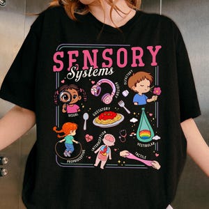 Könnte beinhalten: Schwarzes T-Shirt mit einer Grafik, die die sensorischen Systeme veranschaulicht. Das Design enthält den Text "SENSORY Systems" in Rosa sowie Illustrationen der visuellen, auditiven, gustatorischen, olfaktorischen, propriozeptiven, interozeptiven, vestibulären und taktilen Sinne.