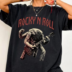 May include: Black t-shirt with the text "ROCKY N ROLL" in red. The shirt features a graphic of a figure playing a guitar. The figure has multiple arms and a textured appearance, giving it a vintage rock and roll vibe.