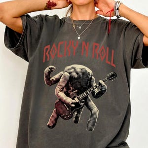 May include: Dark grey t-shirt with the words "ROCKY N ROLL" in red block letters. The shirt features a graphic of a figure playing a guitar. The figure is a light colour with a textured appearance.