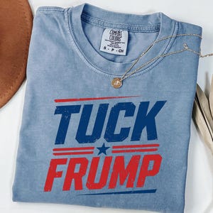 May include: A light blue Comfort Colors t-shirt with the words "TUCK FRUMP" in distressed red and blue lettering. A gold necklace with a pendant is draped across the shirt. The shirt is folded on a white surface.