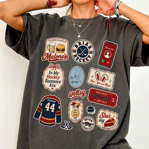 Op de afbeelding: Donkergrijs T-shirt met diverse kleurrijke patches. Patches bevatten tekst als "Malone's", "Briar Hockey", "In My Hockey Romance Era" en "Game Day". Andere patches tonen een hockeytrui, een cocktail en een stuk taart.