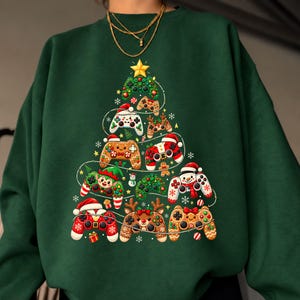 May include: A dark green crewneck sweatshirt featuring a graphic print of a Christmas tree constructed from various video game controllers. The controllers are decorated with holiday themes including Santa hats, reindeer antlers, and gingerbread patterns.