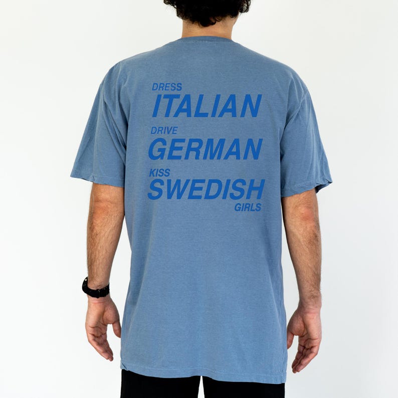 以下が含まれることがあります： 水色のTシャツ。青色の文字で「DRESS ITALIAN DRIVE GERMAN KISS SWEDISH GIRLS」とプリントされています。半袖でカジュアルなスタイルです。
