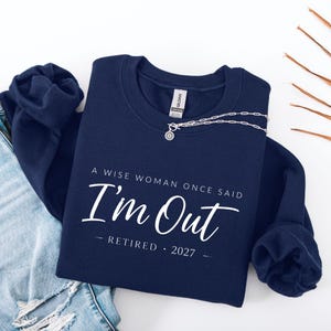 May include: A navy blue crewneck sweatshirt featuring white text that reads A WISE WOMAN ONCE SAID I'm Out RETIRED 2027. The garment has long sleeves and a ribbed collar, with a silver-toned chain necklace resting on the neckline of the fabric.