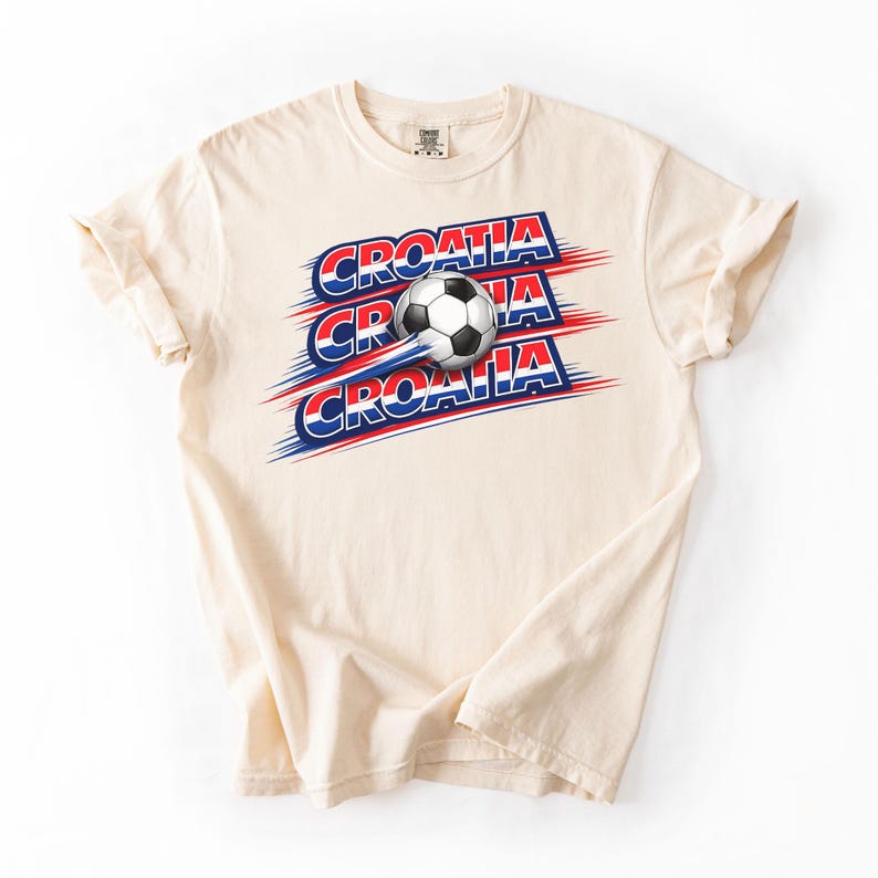 May include: Cream-colored t-shirt featuring the word "CROATIA" in red, white, and blue, with a soccer ball graphic. The text and ball have a dynamic, sporty design, suggesting movement. The shirt is made of soft material.