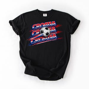 May include: Black t-shirt featuring the word "CROATIA" in red, white, and blue, with a soccer ball graphic. The design includes a soccer ball in motion, with the word "CROATIA" repeated three times.