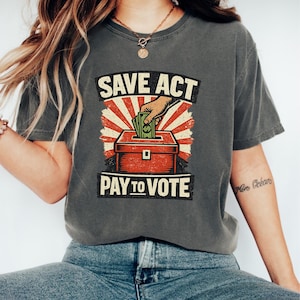 Peut inclure: T-shirt gris foncé avec un motif graphique. Le motif présente le texte "SAVE ACT PAY TO VOTE" au-dessus et en dessous d'une urne rouge avec une main insérant de l'argent. Le fond est rouge et crème.
