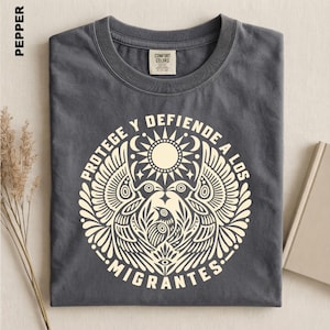 May include: A pepper-colored crew neck t-shirt featuring a cream-colored graphic print of a stylized sun, wings, and a bird. The text "PROTEGE Y DEFIENDE A LOS MIGRANTES" is printed in a circular arrangement around the central bird and wing design.