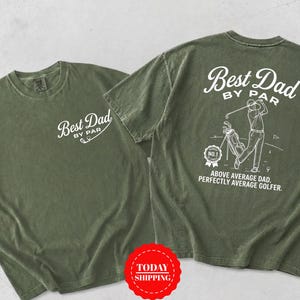 May include: An olive green short-sleeve t-shirt featuring white screen-printed graphics. The front displays the text Best Dad By Par. The back shows a golfer, a golf bag, a No. 1 badge, and the text Best Dad By Par Above Average Dad Perfectly Average Golfer.