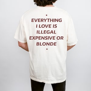 Op de afbeelding: Crèmekleurig T-shirt met de tekst "EVERYTHING I LOVE IS ILLEGAL EXPENSIVE OR BLONDE" in donkerrood. Het shirt heeft een relaxte, oversized pasvorm.