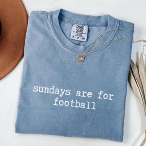May include: A blue Comfort Colors t-shirt with the text "sundays are for football" in white. A gold necklace with a pendant is draped across the shirt. The shirt is size small.