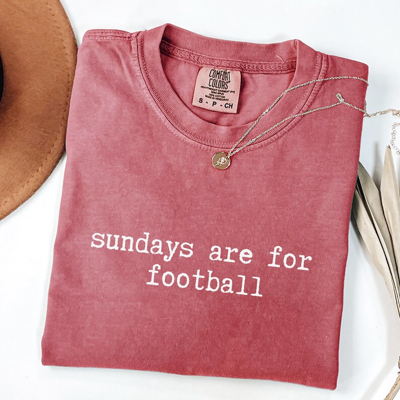 May include: A dusty rose Comfort Colors t-shirt with the phrase "sundays are for football" in white block letters. A silver necklace with a small pendant rests on the shirt. A brown hat is visible in the upper left corner.