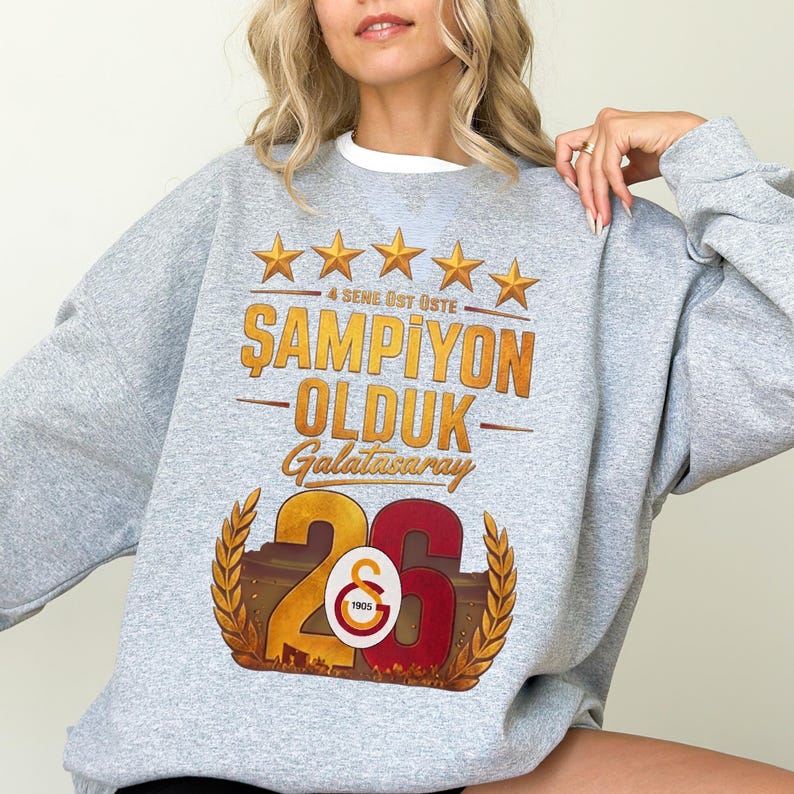 Puede incluir: Sudadera gris con cuello blanco, con texto dorado y estrellas. El texto dice "ŞAMPİYON OLDUK Galatasaray 26". El dise&ntilde;o incluye un escudo con el n&uacute;mero 26 y el logotipo del Galatasaray.