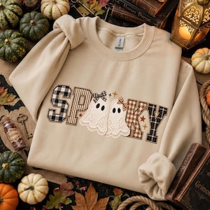 May include: A beige crewneck sweatshirt featuring the word SPOOKY in a patchwork style. The letters are filled with black and white buffalo plaid and brown gingham patterns. Two white ghost illustrations with lace-like edges and small bows are positioned between the letters, accented by small orange stars.