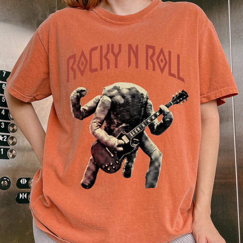 May include: Orange t-shirt with the text "ROCKY N ROLL" in a stylized font. The shirt features a graphic of a figure playing a guitar. The figure is in black and white, contrasting with the orange background.