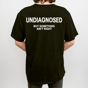 Puede incluir: Camiseta negra con el texto blanco "UNDIAGNOSED" encima de "BUT SOMETHING AIN'T RIGHT". La camiseta es de estilo cuello redondo básico. La imagen muestra la parte trasera de la camiseta.
