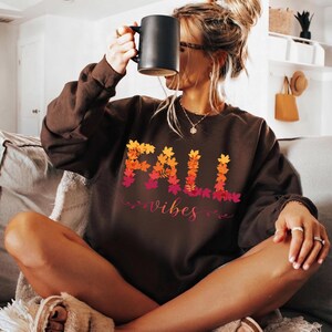 Op de afbeelding: Een donkerbruin sweatshirt met lange mouwen voorzien van de tekst FALL in een grafisch ontwerp van oranje, gele en rode herfstbladeren, met het woord vibes eronder in een cursief lettertype in een bijpassend kleurverloop.