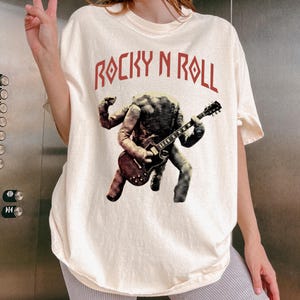 May include: A cream-coloured t-shirt with the text "ROCKY N ROLL" in red. The graphic shows a figure playing a guitar in shades of grey and brown. The t-shirt is a casual style.