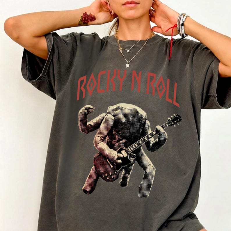May include: Dark grey t-shirt with the words "ROCKY N ROLL" in red block letters. The shirt features a graphic of a figure playing a guitar. The figure is a light colour with a textured appearance.