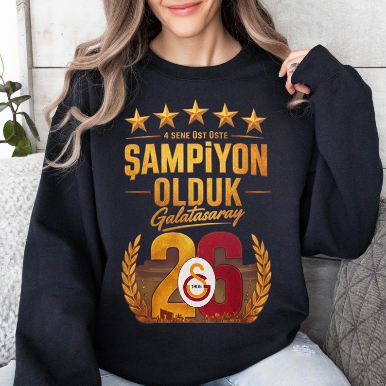 Puede incluir: Sudadera negra con texto dorado y detalles de estrellas. El texto dice "4 SENE &Uuml;ST &Uuml;STE ŞAMPİYON OLDUK Galatasaray 26". El n&uacute;mero 26 es dorado y rojo con un dise&ntilde;o de corona de laurel. La sudadera es de estilo casual y c&oacute;modo.