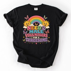 May include: A black t-shirt featuring a colorful graphic print with a rainbow, two smiling suns, and several butterflies. The text reads NEED 2 MALE VOLUNTEERS FOR A THREESOME in bold, multi-colored letters, surrounded by small stars and hearts.