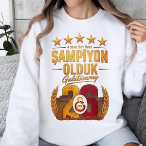 Puede incluir: Sudadera blanca con un dise&ntilde;o gr&aacute;fico. El dise&ntilde;o incluye estrellas doradas, las palabras "4 SENE &Uuml;ST &Uuml;STE", "ŞAMPİYON OLDUK" y "Galatasaray" en oro y rojo. Tambi&eacute;n aparece el n&uacute;mero "26".