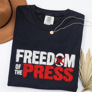May include: A navy blue t-shirt featuring the white text "FREEDOM OF THE" and the red text "PRESS" with a red "X" over the letter "O". The shirt has a Comfort Colors brand label at the neck and is displayed with a gold necklace, a brown hat, and a palm leaf.