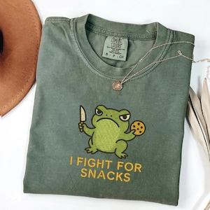 Puede incluir: Camiseta verde Comfort Colors con un dibujo de una rana sosteniendo un cuchillo y una galleta. El texto "I FIGHT FOR SNACKS" está impreso en amarillo. Un collar dorado con un colgante está sobre la camiseta.