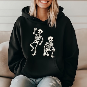 May include: Black hooded sweatshirt featuring a white skeleton graphic. The graphic depicts two skeletons, one waving and the other squatting. The sweatshirt has a front pocket and a drawstring hood, perfect for Halloween or spooky season.