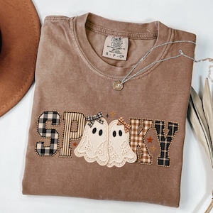 Puede incluir: Una camiseta marrón de manga corta con una etiqueta Comfort Colors en el cuello. El frente presenta la palabra SPOOKY en letras de tela a cuadros y rayas, con dos fantasmas de encaje blanco con lazos pequeños reemplazando la letra O.