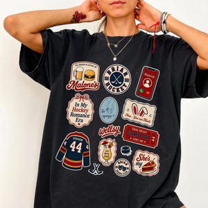 Op de afbeelding: Donkergrijs t-shirt met meerdere grafische patches. Patches bevatten tekst en afbeeldingen gerelateerd aan hockey, eten en zinnen als "In My Hockey Romance Era" en "Game Day". Het shirt heeft een vintage, versleten uitstraling.