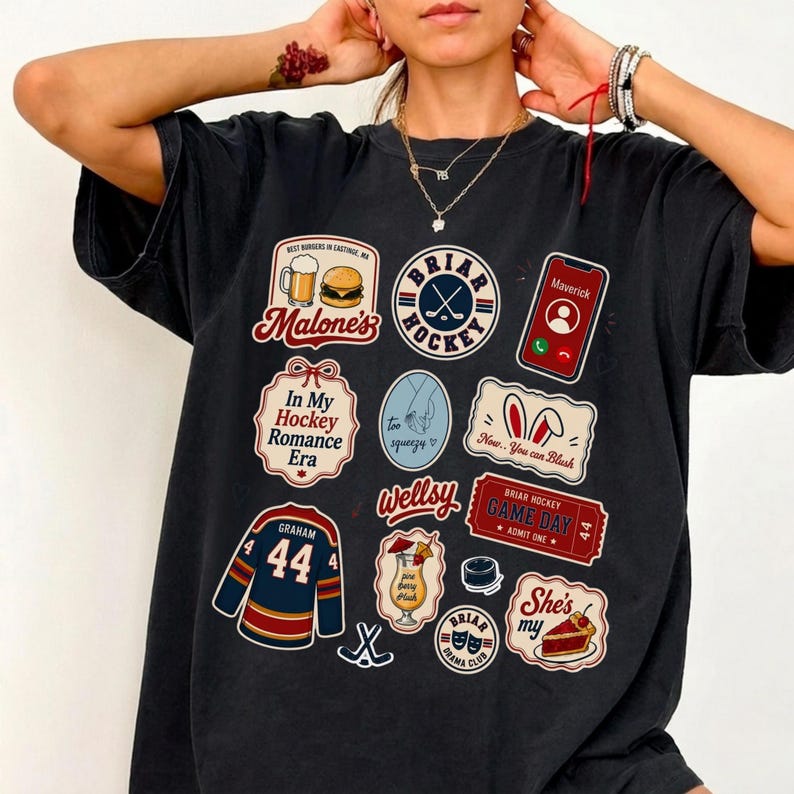 Op de afbeelding: Donkergrijs t-shirt met meerdere grafische patches. Patches bevatten tekst en afbeeldingen gerelateerd aan hockey, eten en zinnen als "In My Hockey Romance Era" en "Game Day". Het shirt heeft een vintage, versleten uitstraling.