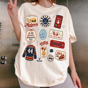 Op de afbeelding: Cr&egrave;mekleurig T-shirt met diverse grafische patches. Patches zijn onder meer een hockeytrui, een telefoon met een oproep en de tekst "Briar Hockey" en "In My Hockey Romance Era".