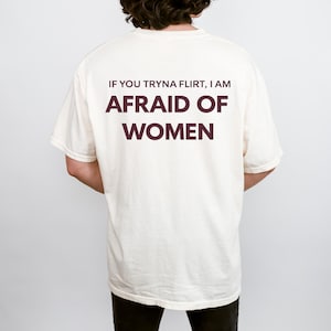 May include: Cream-colored t-shirt with the text "IF YOU TRYNA FLIRT, I AM AFRAID OF WOMEN" in dark burgundy lettering. The shirt is a casual style, with short sleeves and a relaxed fit.