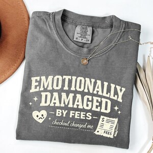 Puede incluir: Una camiseta gris Comfort Colors con el texto "EMOTIONALLY DAMAGED - BY FEES - checkout changed me". La camiseta tiene cuello redondo y mangas cortas. Un collar plateado con un colgante está sobre la camiseta.
